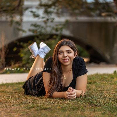 Smiling high school senior girl in a black dress laying on the grass with feet up, posing in front of a stone bridge during golden hour, photographed by Mirage Photography in Michigan.