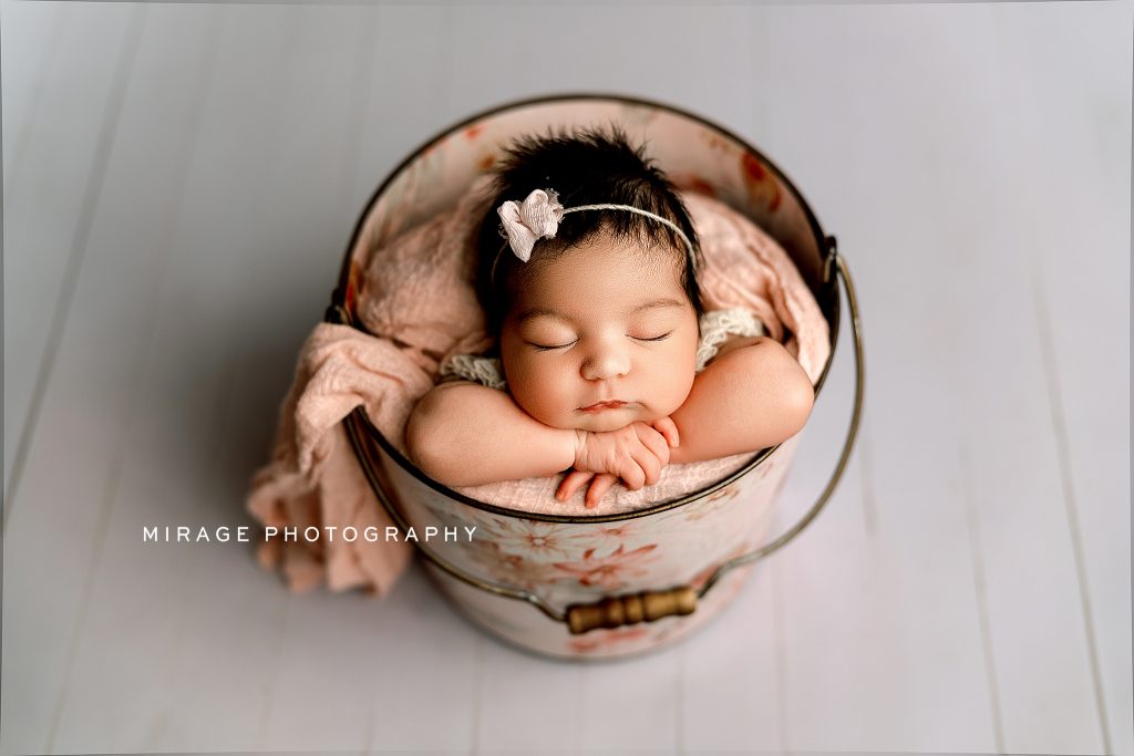 Newborn baby girl with dark hair peacefully sleeping in a floral bucket, posed with arms folded under her chin, wearing a delicate headband and wrapped in soft pink blankets. Studio session by Mirage Photography in Fowlerville, Michigan.