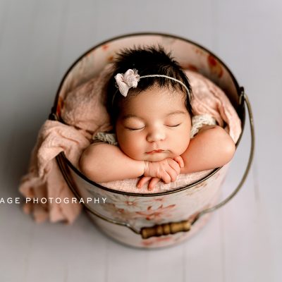 Newborn baby girl with dark hair peacefully sleeping in a floral bucket, posed with arms folded under her chin, wearing a delicate headband and wrapped in soft pink blankets. Studio session by Mirage Photography in Fowlerville, Michigan.
