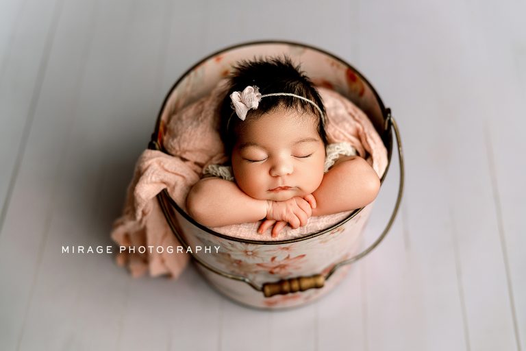 Newborn baby girl with dark hair peacefully sleeping in a floral bucket, posed with arms folded under her chin, wearing a delicate headband and wrapped in soft pink blankets. Studio session by Mirage Photography in Fowlerville, Michigan.