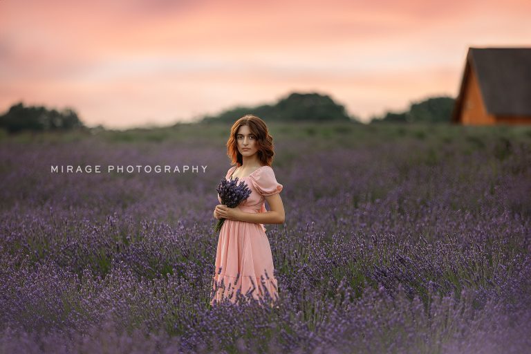 Teen girl in a pink dress holding lavender bouquet in a blooming lavender field at sunset near Fowlerville, Michigan, photographed by Mirage Photography.