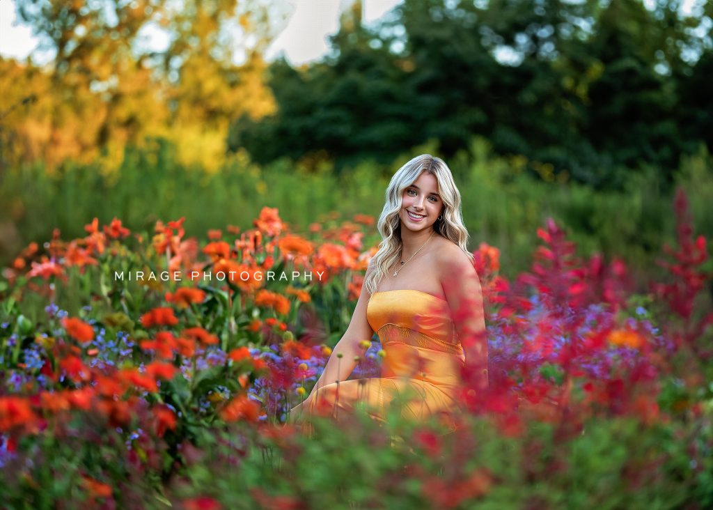 Smiling blonde high school senior in a strapless yellow dress sitting in a vibrant wildflower field during summer, photographed by Mirage Photography in Michigan.