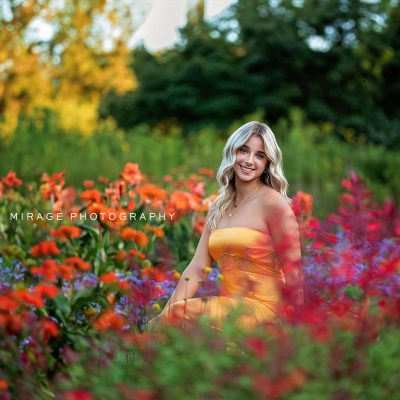 Smiling blonde high school senior in a strapless yellow dress sitting in a vibrant wildflower field during summer, photographed by Mirage Photography in Michigan.