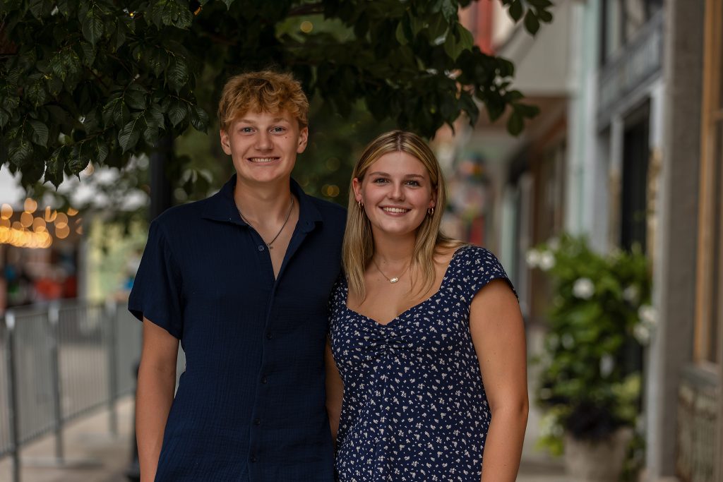 Smiling teen siblings posing together under a tree-lined street in a downtown setting, wearing coordinated navy outfits, captured by Mirage Photography in Michigan.