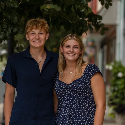 Smiling teen siblings posing together under a tree-lined street in a downtown setting, wearing coordinated navy outfits, captured by Mirage Photography in Michigan.