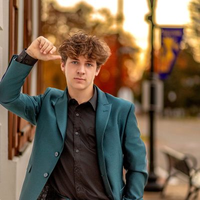 Confident high school senior wearing a teal suit and black shirt, leaning against a downtown building at golden hour in Fowlerville, Michigan, photographed by Mirage Photography.