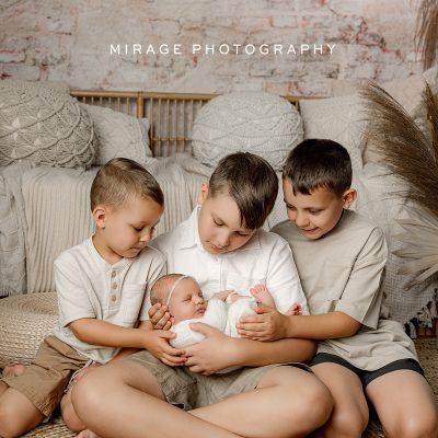 Three older brothers gently holding their newborn baby sibling during a lifestyle studio session by Mirage Photography. Neutral tones and cozy textures in the background.