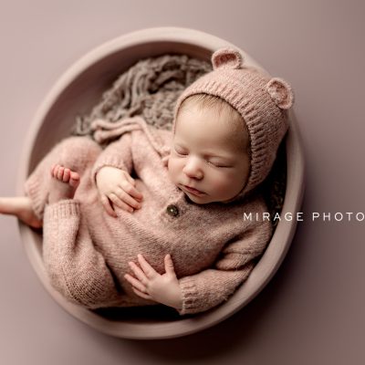 Newborn baby wrapped in a cream knit blanket and hat, peacefully sleeping while holding a tiny stuffed animal, photographed by Mirage Photography in a rustic wood setting.