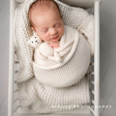Newborn baby swaddled in white knit wrap holding a small teddy bear, photographed in a white vintage crib by Mirage Photography in Fowlerville, Michigan