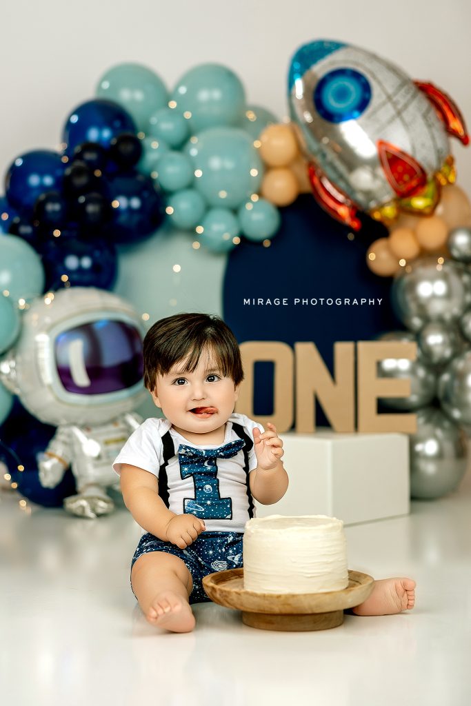 One-year-old boy celebrating his space-themed cake smash session with a small white cake in front of him, surrounded by galaxy-themed balloon garland, astronaut and rocket balloons, and a "ONE" sign in the background.