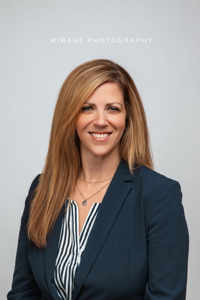 Professional studio headshot of a smiling businesswoman in a navy blazer, taken at Mirage Photography in Fowlerville, Michigan.