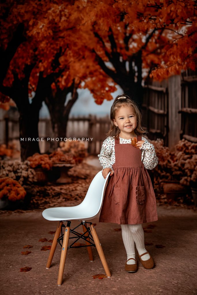 Preschool girl in a fall-themed school photo session, smiling in front of autumn leaves backdrop, holding a leaf — Mirage Photography, Fowlerville MI