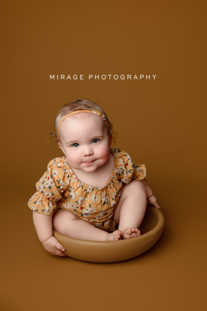 Baby girl in floral romper sitting in a neutral bowl during milestone photography session – Mirage Photography, Fowlerville MI