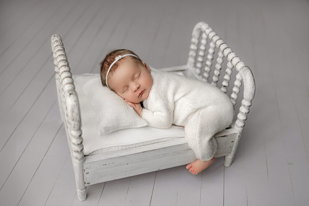 Sleeping newborn baby in a soft white knit outfit and headband, posed on a small vintage white bed with matching pillow and blanket during an all-white studio newborn session at Mirage Photography in Fowlerville, Michigan.