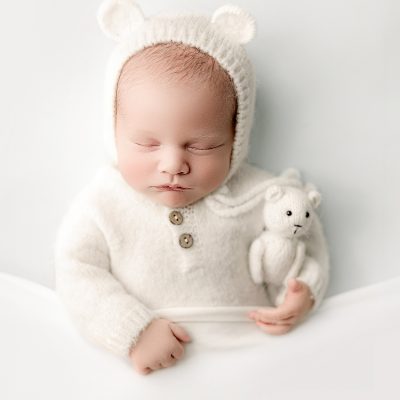 Newborn baby boy in soft white bear bonnet and sweater holding a teddy during a studio session with Mirage Photography in Fowlerville, Michigan