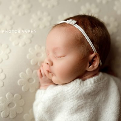 Newborn baby girl sleeping on floral cream blanket wearing white knit outfit and bow headband photo by Mirage Photography in Fowlerville, MI