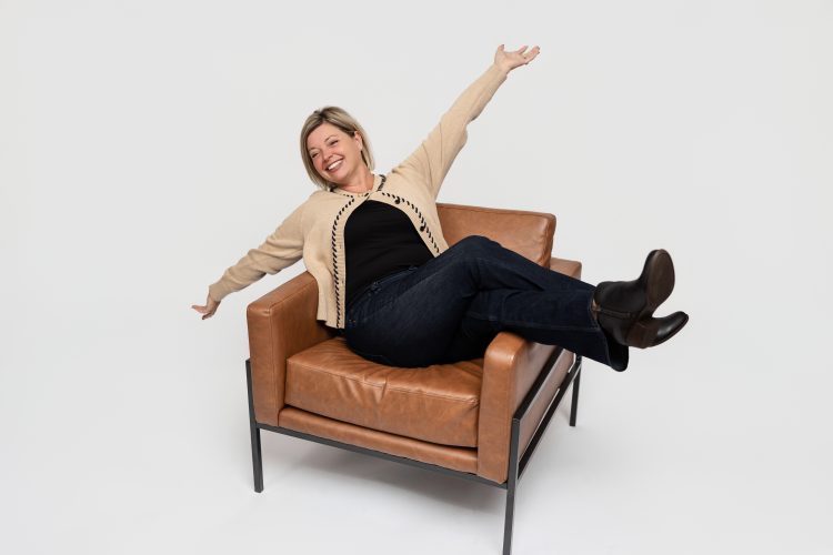 Playful personal branding headshot of smiling professional woman sitting in a modern leather chair with arms outstretched, wearing a beige cardigan, black top, and jeans – Mirage Photography Fowlerville Michigan.
