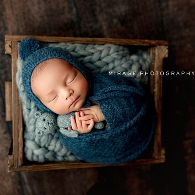 Newborn baby wrapped in deep blue knit swaddle sleeping in a wooden crate during a cozy studio session at Mirage Photography in Fowlerville Michigan